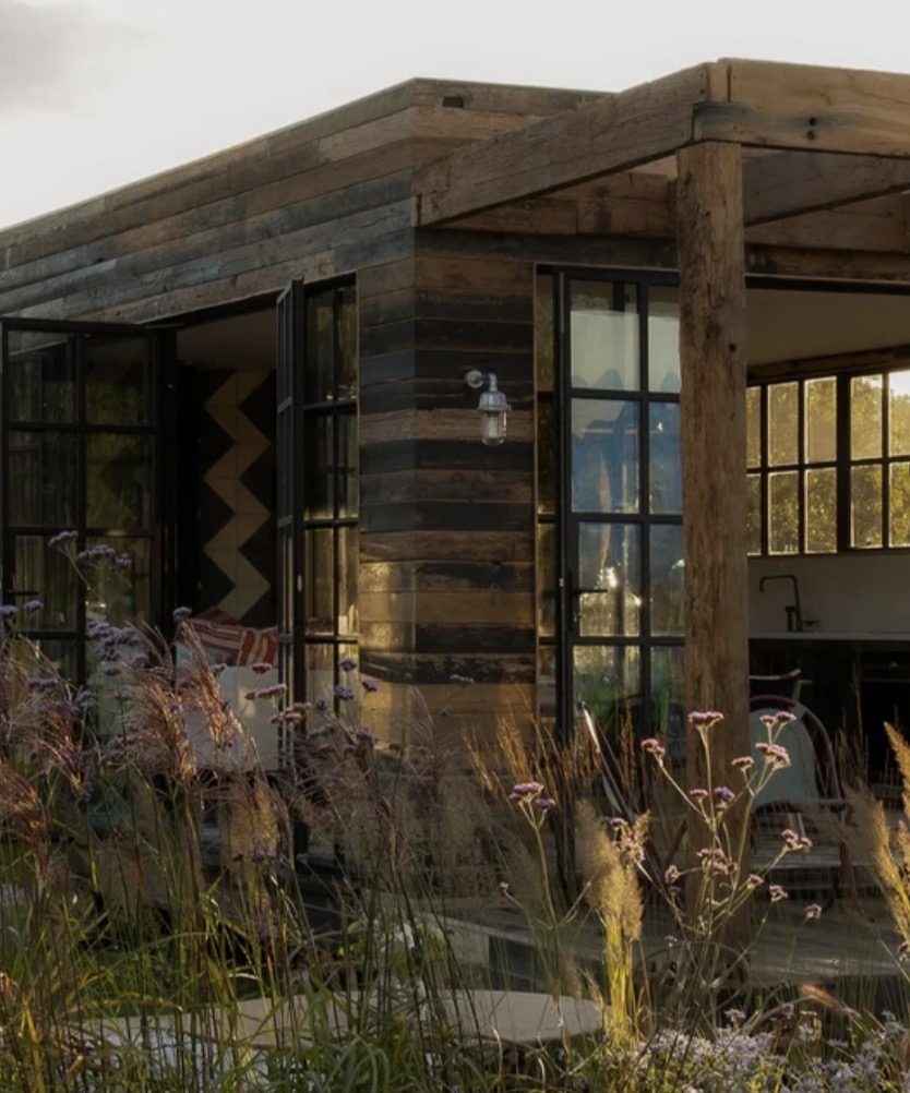 A BERTS cabin at The Pig, New Forest: reclaimed-timber cladding, steel-framed glazing and a timber pergola, among tall grasses and verbena at golden hour
